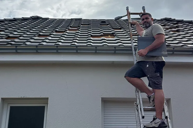 Jonathan, dirigeant de SJ Rénovation, sur une échelle contre une toiture en tuiles, une tuile à la main